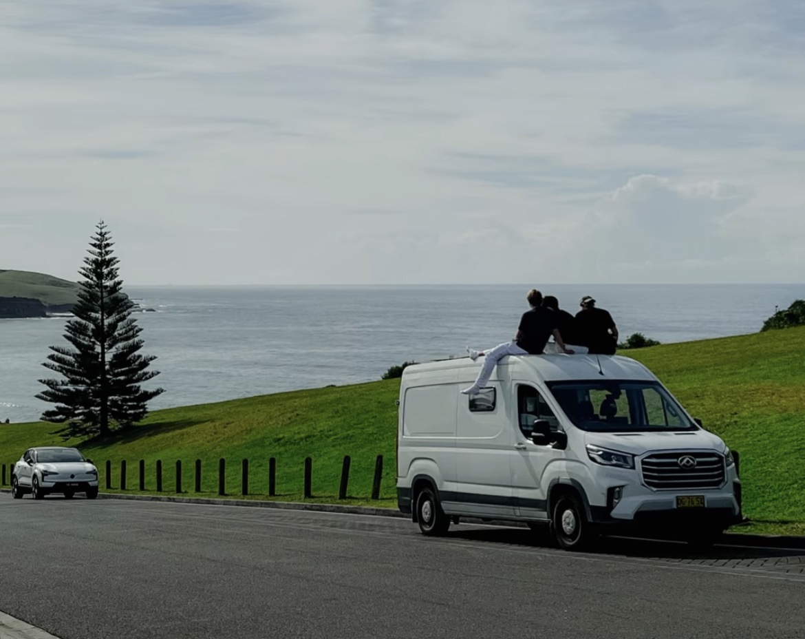 A white campervan parked on a grassy headland above the ocean, travellers on the roof